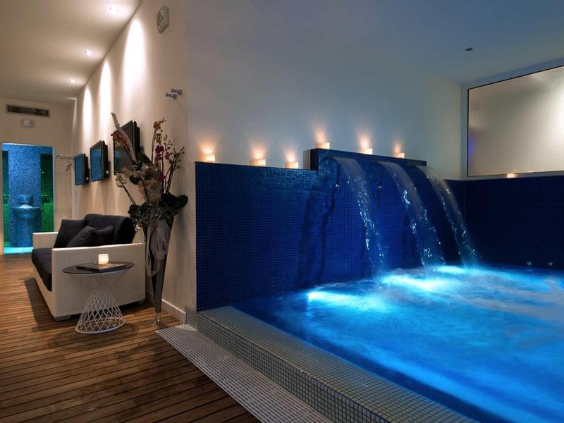 Moderner Wellnessbereich mit beleuchtetem Innenpool, Wasserfall und gemütlichem Sitzbereich.