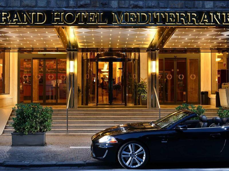 Eingang des Grand Hotel Mediterraneo mit goldfarbenen Lichtern und schwarzem Cabrio vor der Tür.