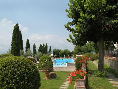 Area piscina con lettini e giardino curato con cipressi e alberi all'ombra sotto il sole.