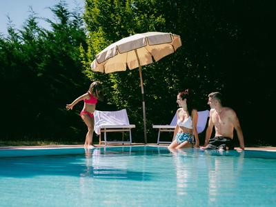 Tři lidé si užívají slunečný den u bazénu s slunečníkem a lehátky.