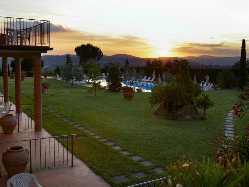Západ slunce nad hotelovou zahradou s bazénem a kopci v pozadí.