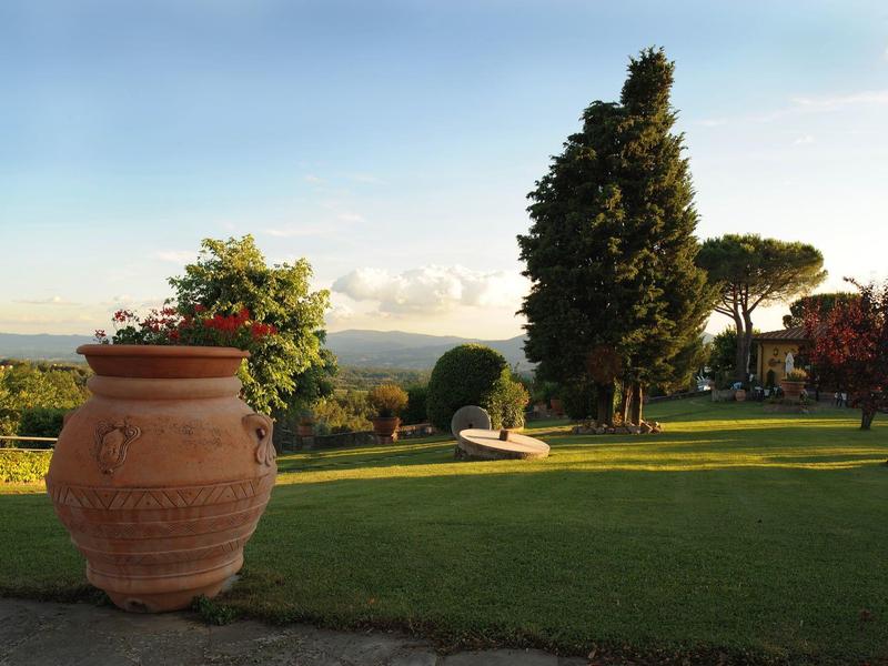 Velký terakotový květináč na upraveném trávníku s stromy a vzdáleným výhledem.