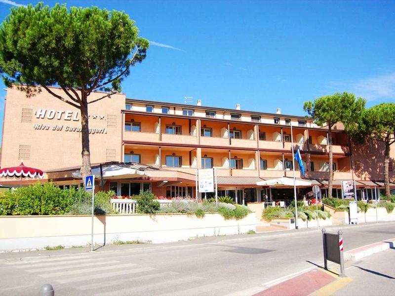 Edificio di hotel a tre piani con area esterna e ombrelloni lungo una strada trafficata.
