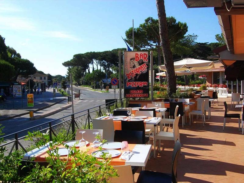 Area esterna di un caffè con tavoli apparecchiati e ombrelloni lungo una strada tranquilla.