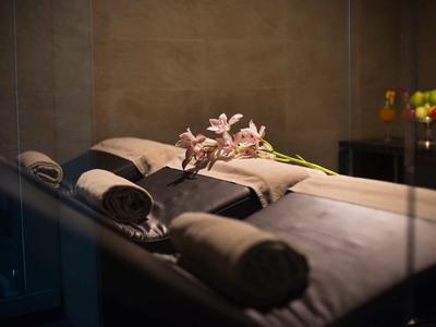 Ein ruhiger Wellnessraum mit einer Massageliege, grauen Handtüchern und rosa Blumen darauf.