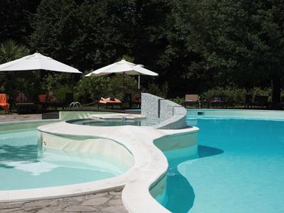 Zwei verbundene Schwimmbecken mit weißen Sonnenschirmen und grünen Bäumen im Hintergrund.