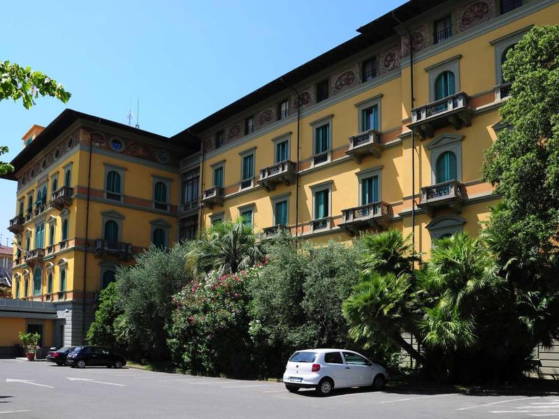 Gelbes Wohnhaus mit Balkon und grünen Büschen im Hof, blauer Himmel, weißes Auto auf Parkplatz.
