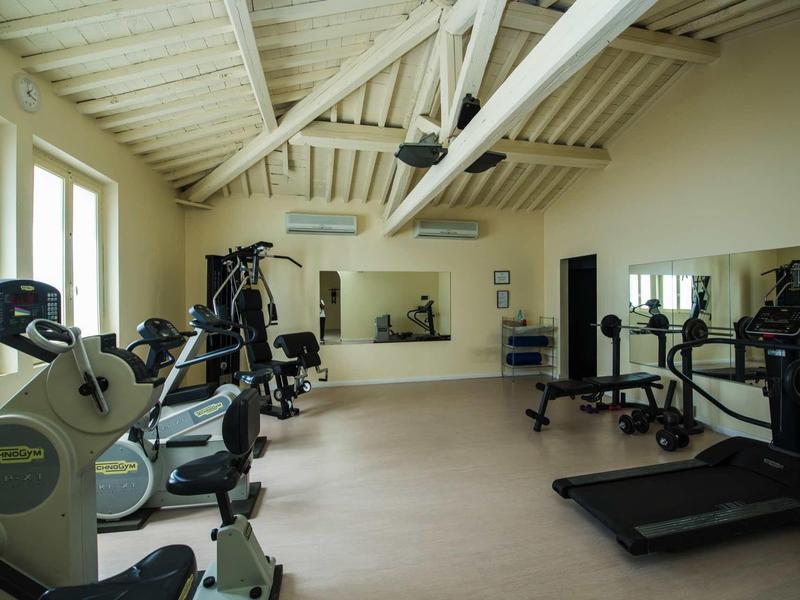 Heller Fitnessraum mit weißen Holzbalken, mehreren Sportgeräten und großen Fenstern.