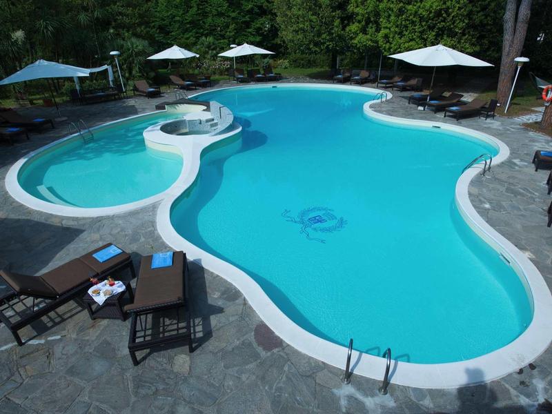 Schwimmbecken mit unregelmäßiger Form, blauer Wasserfarbe, umgeben von Liegestühlen und Sonnenschirmen.