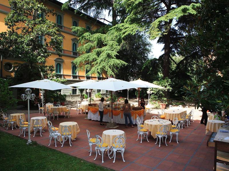 Außenterrasse mit weißen Tischen und Stühlen, Sonnenschirmen, umgeben von Bäumen und Gebäude.