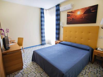 Camera d'hotel con testata gialla, copriletto blu, mobili in legno e luce diurna attraverso le tende.