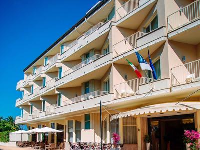 Großes Hotelgebäude mit Balkonen, Flaggen, Sonnenschirmen und Straßencafé bei blauem Himmel.