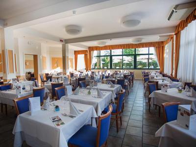 Jasna restauracja z białymi obrusami, niebieskimi krzesłami i dużymi oknami z widokiem na zieleń.