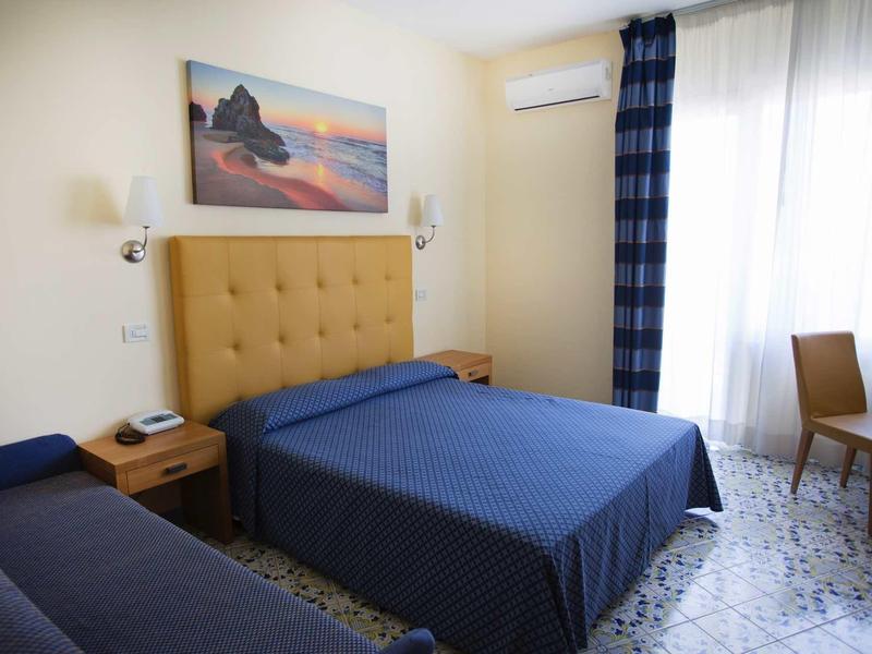 Camera d'hotel con letto matrimoniale, divano, testiera gialla e quadro alle pareti.