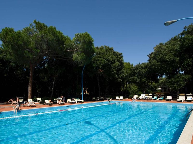 Grande piscina all'aperto con acqua azzurra limpida, circondata da lettini e alberi sotto un cielo limpido.