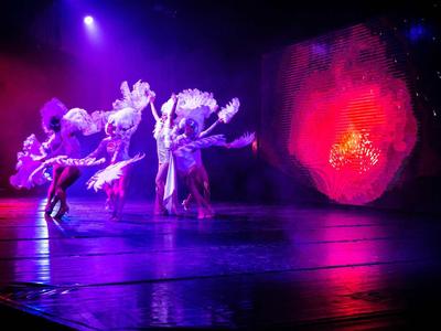 Ballerini in costumi colorati su un palco con luci rosa-viola e una proiezione rossa sullo sfondo.