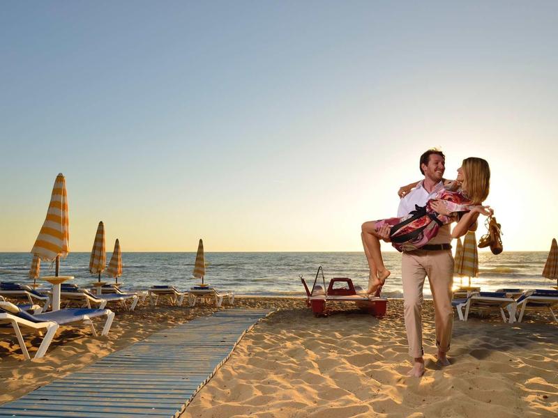 Persona che porta un bambino sulla spiaggia al tramonto con lettini e ombrelloni.