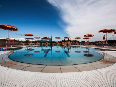 Kruhový bazén s červenými slunečníky a lehátky pod jasnou oblohou.