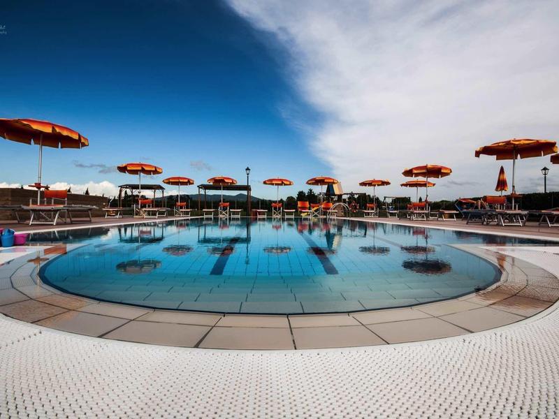 Kruhový bazén s červenými slunečníky a lehátky pod jasnou oblohou.