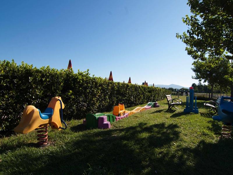 Venkovní hřiště s barevnými pružinovými houpačkami a keři pod jasnou oblohou.
