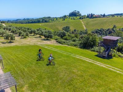 Dva cyklisté jedou přes zelenou louku s mírně zvlněnými kopci v pozadí.