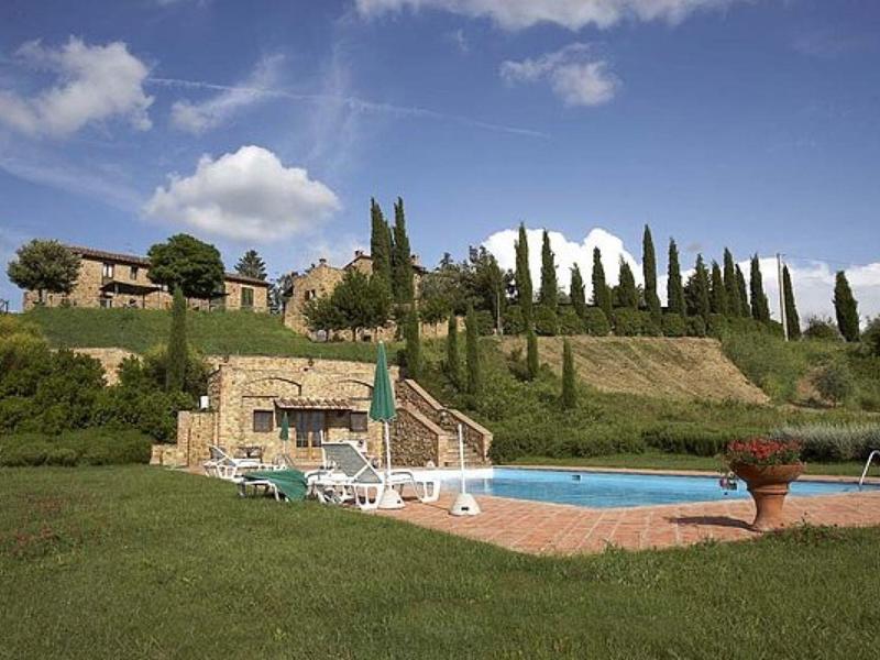 Bazén s lehátky před kopcem s cypřiši a budovami pod modrou oblohou.