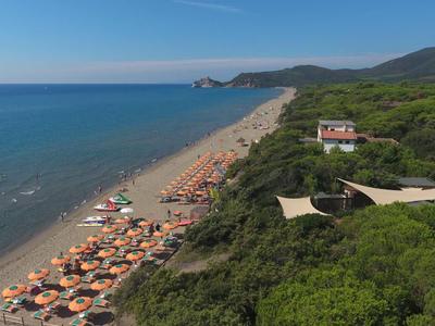 Langer Sandstrand mit Sonnenschirmen und klarem Meer neben grün bewaldetem Hügel und Häusern.