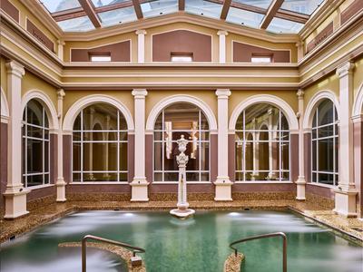 Nasłonecznione patio hotelowe z basenem i klasyczną statuą pod szklanym dachem.