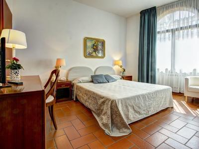 Jasny pokój hotelowy z podwójnym łóżkiem, drewnianymi meblami i dużymi oknami z zasłonami.