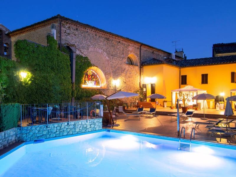 Oświetlony basen i strefa wypoczynkowa przy historycznych budynkach nocą.