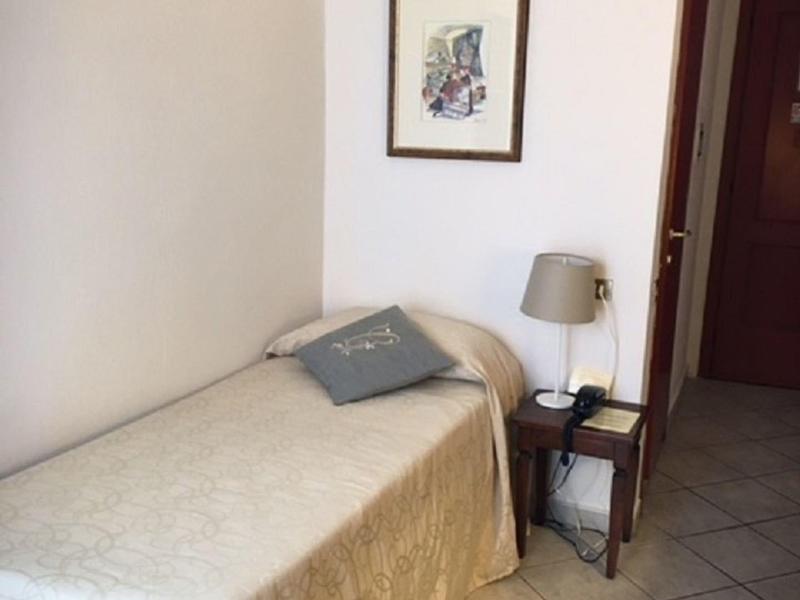 Łóżko pojedyncze z beżową narzutą, poduszką i szafką nocną z lampką obok białej ściany.