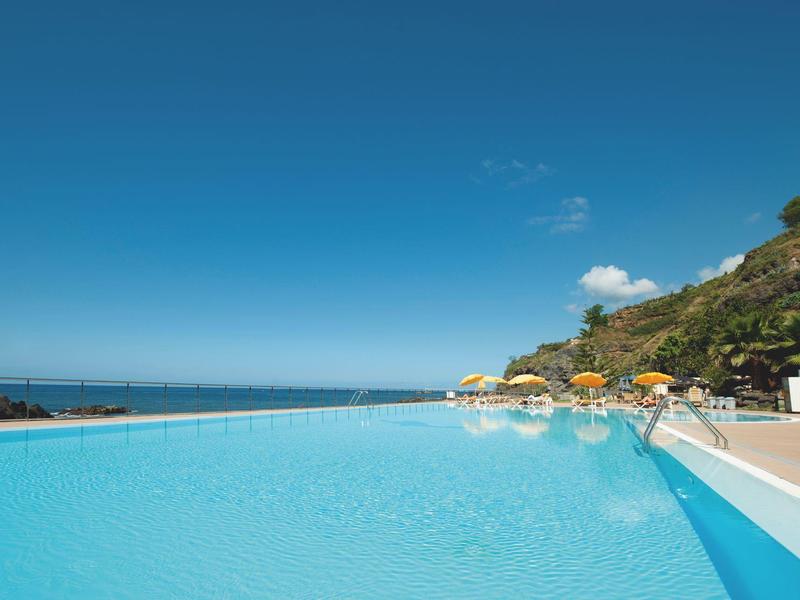 Piscine à débordement avec vue sur la mer calme et transats avec parasols près d'une colline rocheuse.