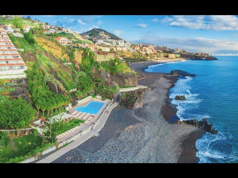 Complexe de luxe sur des falaises verdoyantes donnant sur une plage de sable noir et une mer bleue sous un ciel nuageux.
