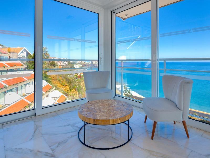 Coin de chambre d'hôtel en bord de mer avec murs en verre, chaises blanches et table ronde en bois