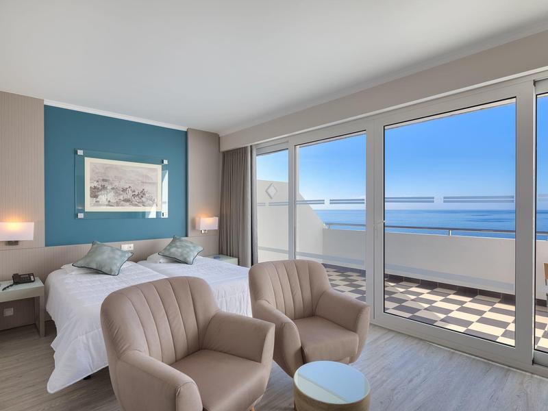 Chambre d'hôtel lumineuse avec vue sur la mer, lit double, deux fauteuils et balcon.