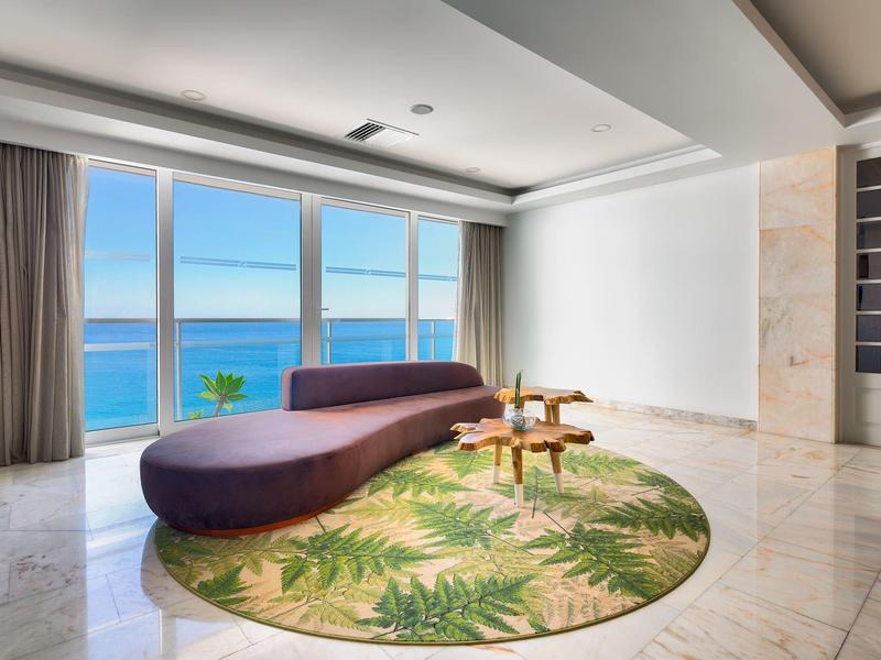 Espace lounge moderne avec canapé violet incurvé, tapis rond à feuilles vertes et vue sur l'océan.