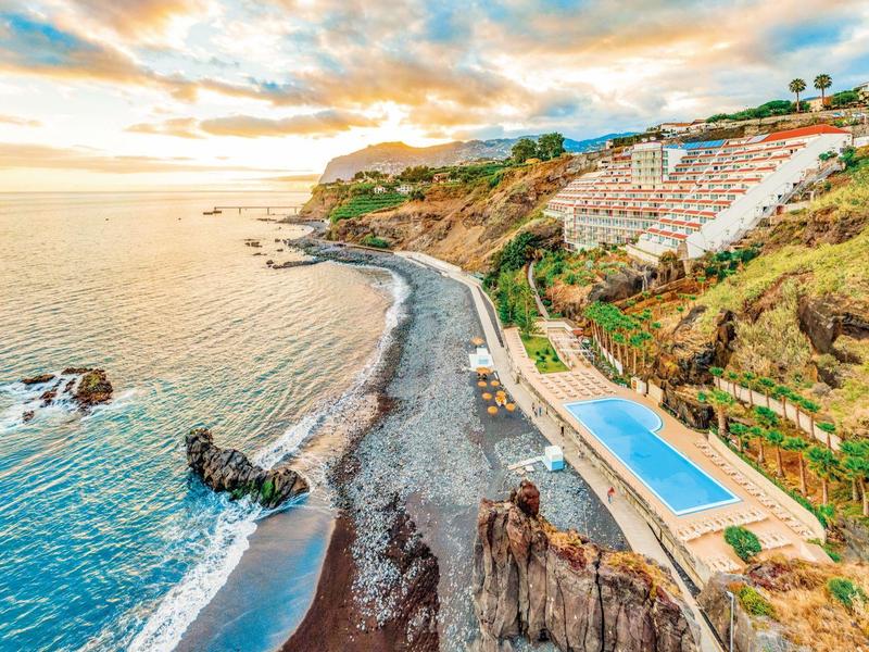 Station balnéaire avec falaises, plage de galets et piscine surplombant l'océan au coucher du soleil.