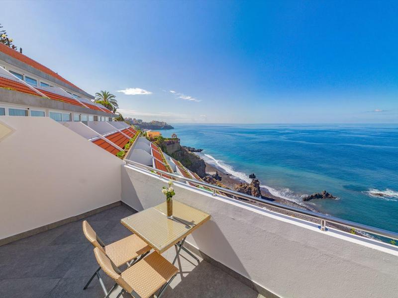 Balcon avec table et chaises donnant sur une côte rocheuse et une mer bleue sous un ciel clair.