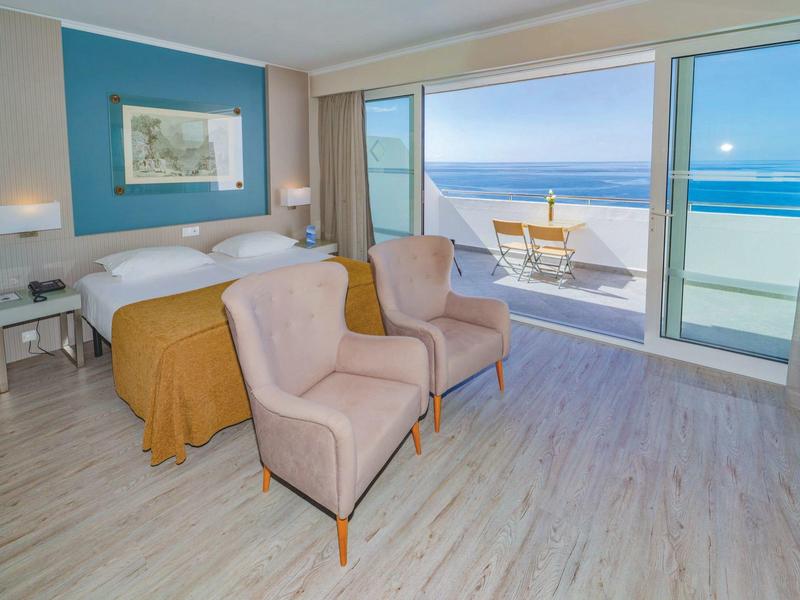 Chambre d'hôtel lumineuse avec vue sur l'océan, lit double, deux fauteuils et balcon.