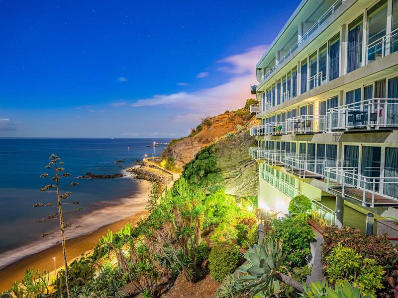 Bâtiment d'hôtel en bord de mer surplombe la plage et l'océan sous un ciel bleu clair au coucher du soleil.