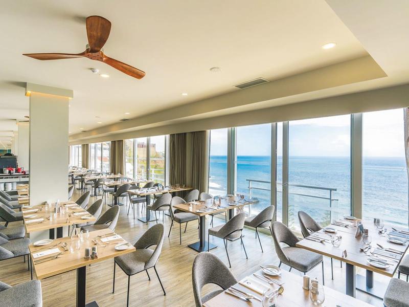 Restaurant lumineux avec vue sur la mer, tables en bois, chaises modernes, ventilateur au plafond.