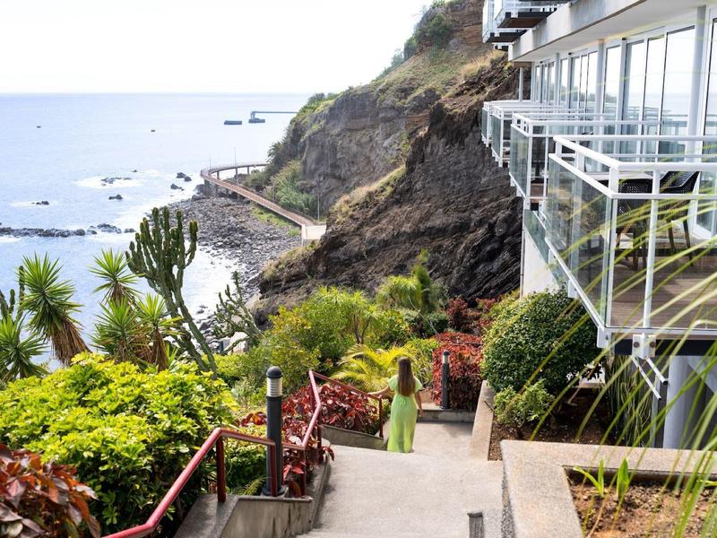 Terrasse d'hôtel en bord de mer avec vue sur l'océan et verdure luxuriante le long d'un chemin en pente.
