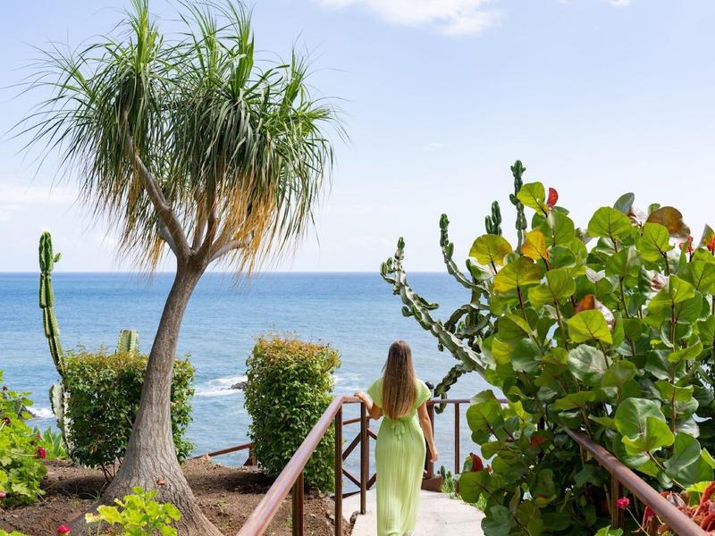 Femme en robe claire descend des marches en bois vers la plage avec vue sur l'océan et plantes tropicales.