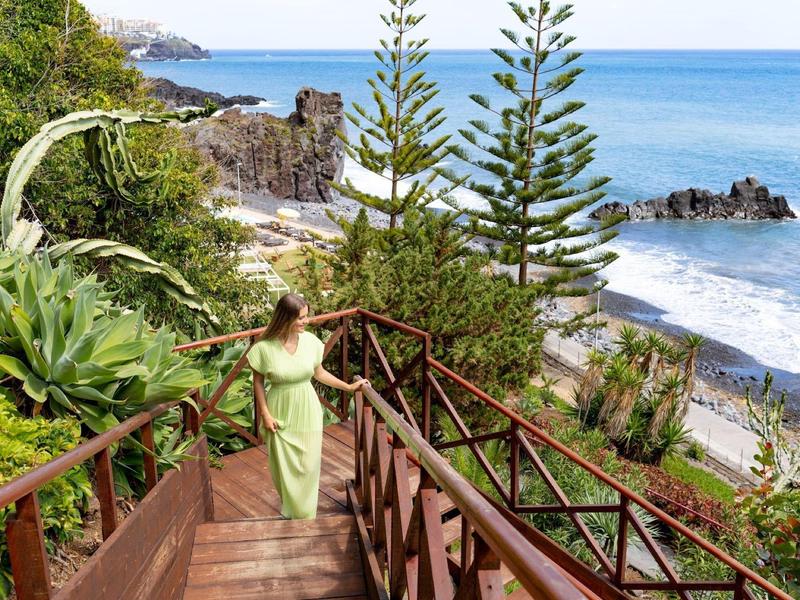 Femme en robe verte descend des escaliers en bois près de la plage avec arbres et vue sur l'océan.