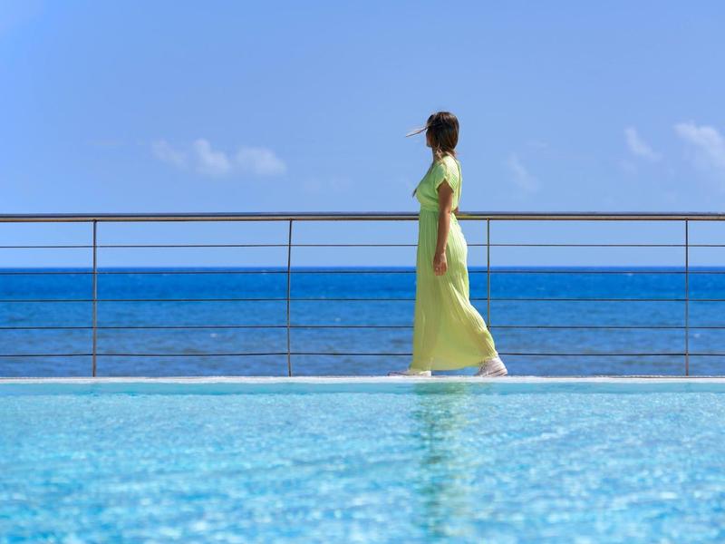 Une personne en robe jaune marche à côté d'une piscine bleue claire avec l'océan et le ciel en arrière-plan.