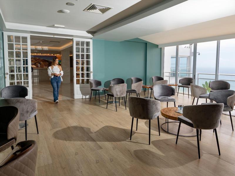 Salon d'hôtel spacieux avec chaises modernes, petites tables, grandes fenêtres et une personne marchant.