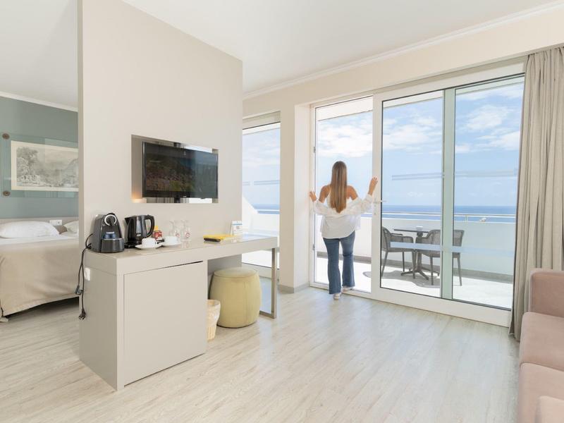 Chambre d'hôtel lumineuse avec balcon, vue sur la mer et femme regardant dehors.