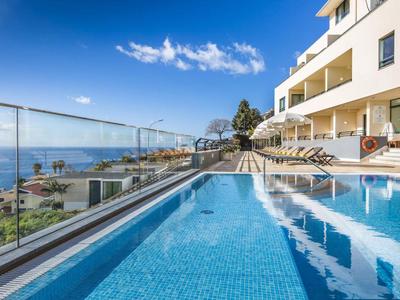Piscina al aire libre junto a un edificio con vistas al mar y tumbonas bajo un cielo azul claro.