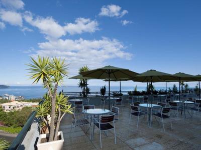 Terraza con mesas y sillas, vista al mar, colinas y ciudad bajo cielo despejado.