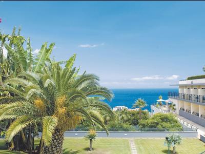 Complejo hotelero con palmeras, flores coloridas y vista al mar azul bajo cielo despejado.